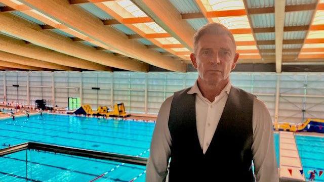 Chris kelly, who has light hair swept over to one side, stands in a waist coat and white shirt. The pool is seen behind him 