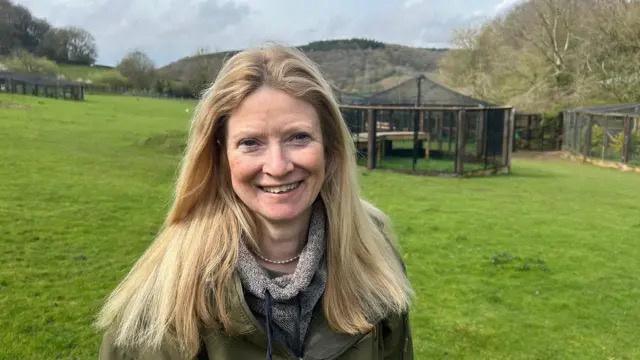 A lady with shoulder length blonde hair is smiling into the camera. She is standing on a large patch of grass with an animal enclosure in the distance behind her. 