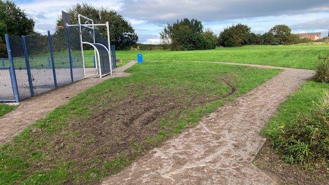 Churned-up grass next to a multi-sports court surrounded by a blue metal fence.