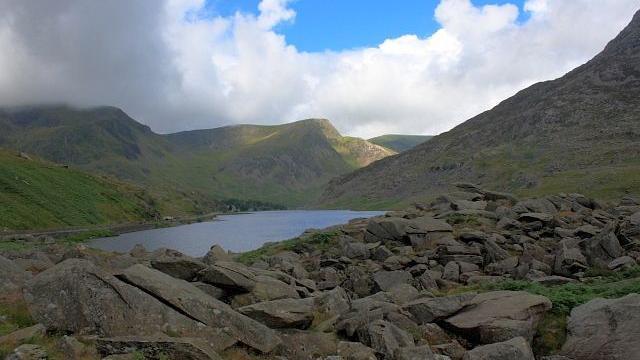 A mound of rocks is shown in front of a small lake with expansive mountains surrounding them.