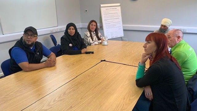 Six people sitting around a table. There are three women and three men in a room with a white board in the far corner of the room