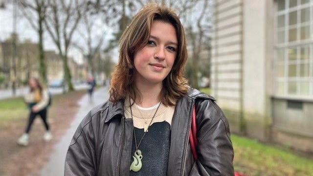 Kristin Millward stands outside. She is wearing a brown leather jacket, cream and grey jumper and a jade necklace.