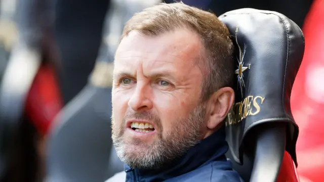 Nathan Jones, wearing a dark blue tracksuit top, grimaces while sitting on the bench at Swansea during Charlton's 3-1 defeat.
