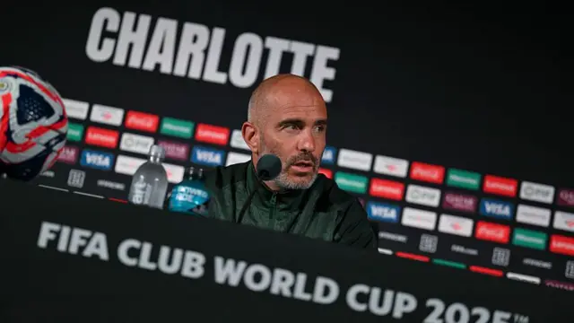 Enzo Mareca sitting behind a table during a news conference with a microphone partially obscuring his face. He is sitting in front of a black background with a variety of sponsors' logos and the word 'Charlotte'. There is a football on the table beside him and 'Fifa Club World Cup 2025 is written on the front of the desk.