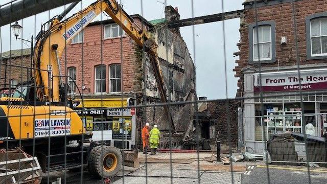 Two workmen in hi-vis clothing inspect the site of the demolished Bakehouse. There are shops on either side and a digger parked on the road.