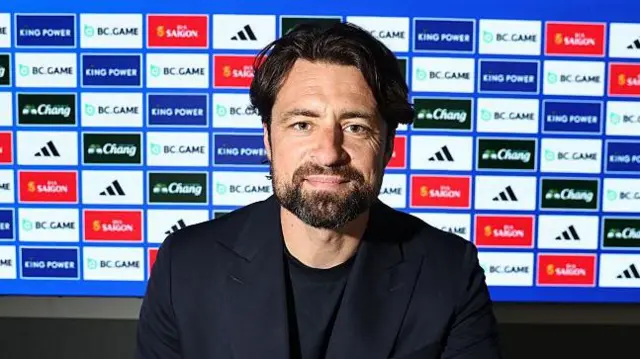 New Leicester City boss Russell Martin sitting in front of a board bearing the club's sponsors while wearing a dark blue suit jacket and dark blue t-shirt