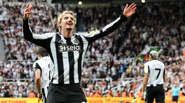 Nick Woltemade, wearing Newcastle black and white home shirt and holding his arms aloft in celebration after scoring a goal. Sandro Tonali and Joelinton are in the background, as is the out of focus crowd at St James' Park.