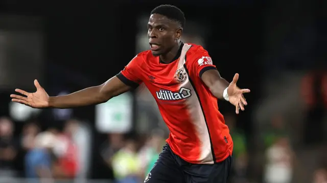 Chiedozie Ogbene holds out his arms as if he is pleading and looks right to left with his mouth partially open. He is wearing Luton's orange home kit with a white stripe down the front left side. 