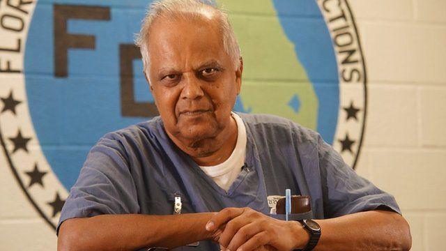 Kris Maharaj in a blue prison uniform with a white t-shirt underneath. He is looking at the camera. Painted on the wall behind him is a large round logo for the Florida Department of Corrections