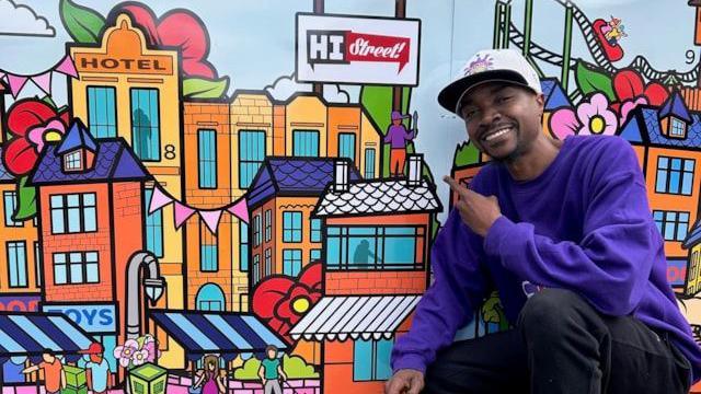 Artist Marvin Mudzongo, in a purple jumper and black jeans, squats next to a colourful digital picture of buildings.
