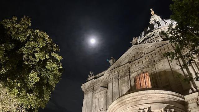 The full moon shines through the clouds over a large stone building with a doomed roof. There are two trees illuminated in the foreground.