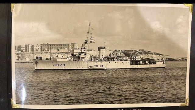 Black-and-white photo of a navy ship on water with buildings in the background.