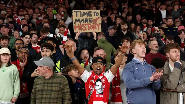 Arsenal fans celebrate in the stands. One holds a sign which reads 'time to make history'