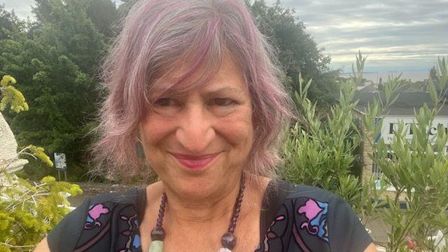 A woman with pink and grey hair lookings into the camera, half-smiling. She wears a beaded necklace and a colourful shirt. Behind her there is some vegetation.