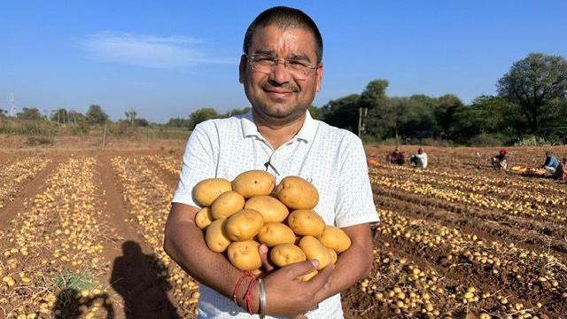 Smiling, Jitesh Patel cradles a pile of potatoes in his arms while standing in a field.