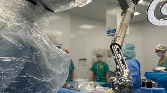 Robotic metallic coloured arm hanging down with surgeons in the background.