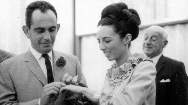 George is pictured on the left, putting a ring on his wife's finger during their wedding. He has short dark hair and wears a light shirt with a dark tie and what looks like a carnation on his lapel. Anna has her hair in a large, elegant updo swept back from her face, and wears a light shirt or jacket with ruffles around her neck. 