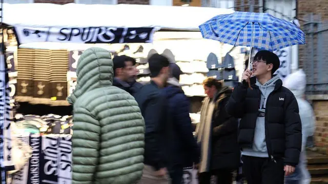 A fan passes a scarf seller with an umbrella ahead of the Premier League match between Tottenham and Manchester City at Tottenham Hotspur Stadium