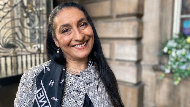 Linda Priddie smiles for the camera, she is wearing a navy blue Chanel scarf and a blazer with a black, white and grey geometric pattern. She has long dark hair and is standing outside Liverpool Town Hall, whose stonework can be seen in the background.