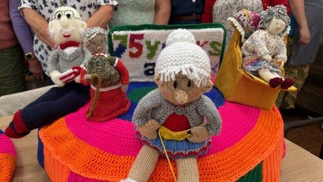 A close-up of a knitted postbox topper. There are four knitted people on there - a man with glasses sitting down, a woman with a walking stick, a woman sitting down and knitting and another woman sitting on a yellow armchair. A knitted sign which is partly visible reads "75 years age uk"