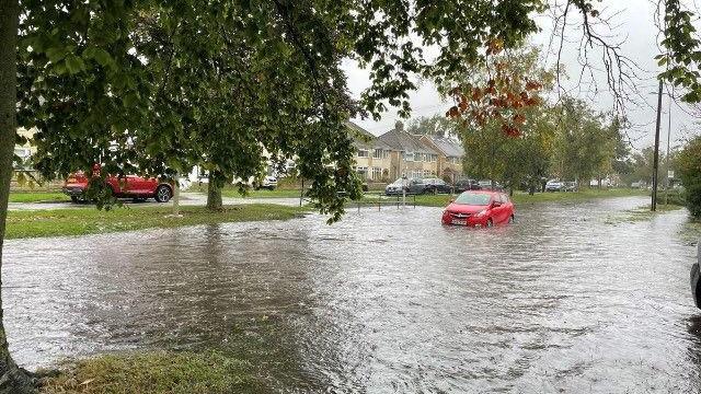 A red car sits in a large area of water, with pavements and houses on either side 