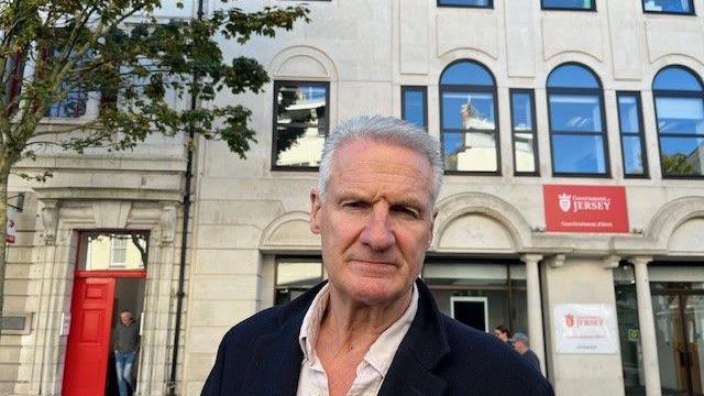 Tom Binet looking at the camera wearing a white shirt and dark blue coat. In the background is a light grey building which has a red sign on the right with the text 'Government of Jersey'. 