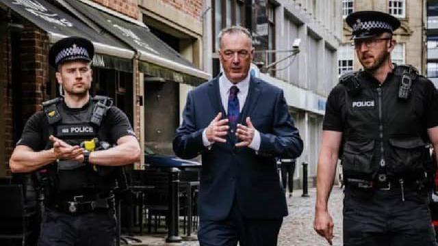 Clive Grunshaw, in the middle, out on patrol with two uniformed police officers. He wears a blue suit, white shirt and blue tie. 
 