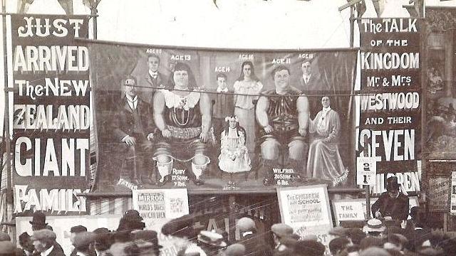 A black and white photo of people in Victorian dress standing around a circus tent with a poster advertising New Zealand's giant family and a picture of nine people - two of which are extremely large.