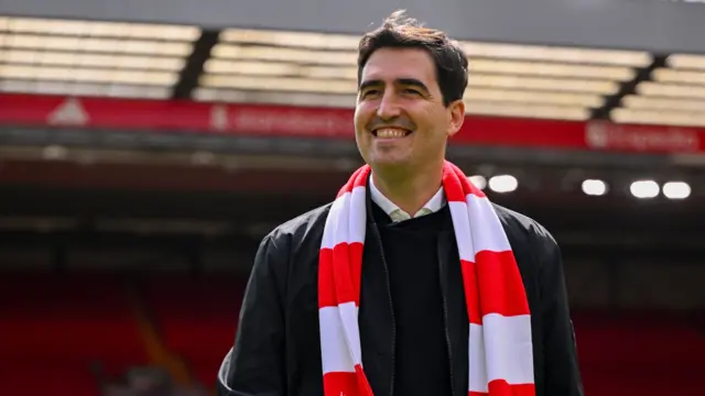 Andoni Iraola smiles while standing on Anfield pitch wearing a Liverpool red and white scarf