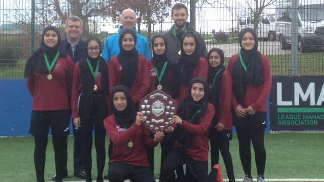 A group picture of the team at a previous tournament. The girls are wearing black headscarves and deep red long-sleeve T-shirts and black shorts and leggings. Two girls are holding up a winning shield. Mr Wright and another coach stands in the background