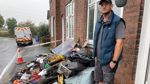 A man wearing a black baseball hat, grey t-shirt, shorts and a blue body warmer is standing next to a pile of burnt belongings.