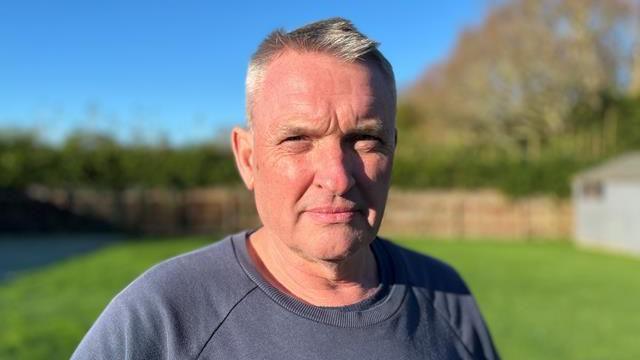 Jon Giles standing in his garden wearing a grey jumper looking at the camera with a furrowed brow. He has grey hair 