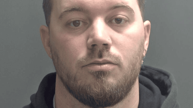 Christopher Rudd is staring at the camera and has a light brown and beard and earrings. He is wearing a dark hoodie and is standing against a plain grey background.