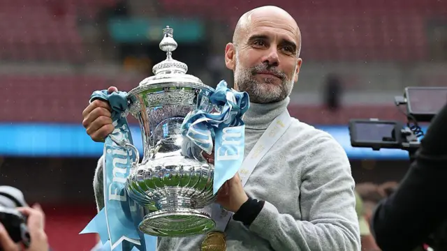 Manchester City's manager Pep Guardiola poses with the FA Cup trophy on the pitch.