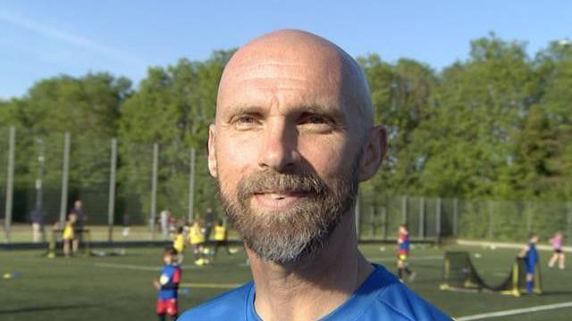 Close-up image of Luke Chadwick. He has a bald head and beard and is smiling at the camera. He is wearing a blue T-shirt. Behind him are some pitches where children are playing football.