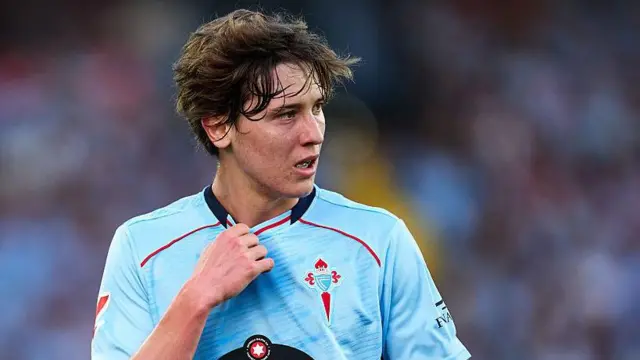 Fer Lopez in action for Celta Vigo. He is wearing a light blue shirt and is holding the neckline with his right hand while looking to his left at the action.