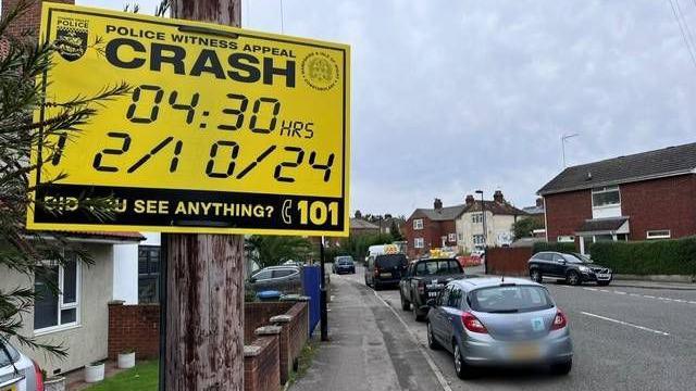 A yellow police witness appeal sign is fixed to a post near a T-junction in a residential area