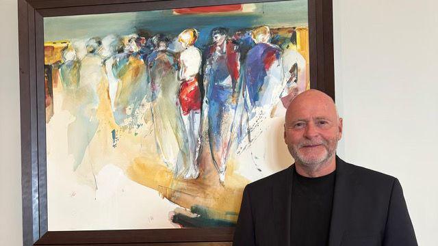 A man in a black suit jacket and black T shirt smiling in front of the camera in front of an artwork