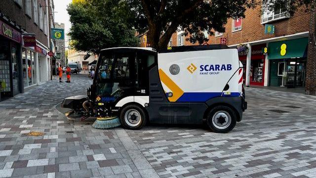 A blue white and yellow street cleaning vehicle, with the word Scarab on the side, scrubs the grey and white paving of the pedestrianised area of St George's Street in Canterbury.