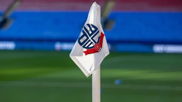 A white corner flag at Bolton Wanderers' Toughsheet Community Stadium with the club's navy blue and red logo on. The pitch and the red and seats of the Toughsheet Community Stadium can be seen blurred out of focus behind.