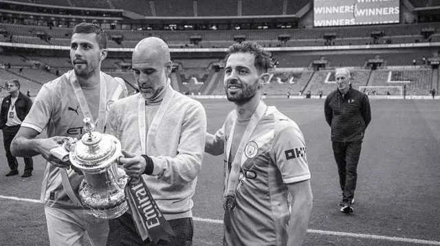 Black and white image of Guardiola holding the FA Cup. Rodri and Bernardo Silva are next to him. They stand on the Wembley pitch.