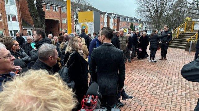 A crowd of people gathered together. Behind them a row of houses is visible