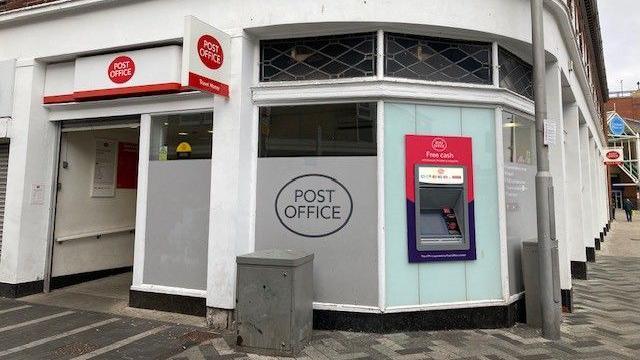 A photo of the exterior of Grimsby Post Office, with a cash machine, entrance door and patterned paving outside.