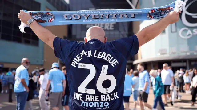 A Coventry City fan holds up a scarf while wearing a replica shirt which reads "Premier League, 26, We Are Back"