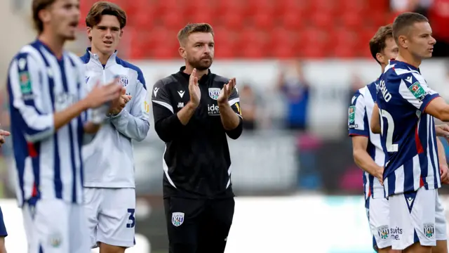 James Morrison, wearing a black West Bromwich Albion tracksuit, stands alongside his players to applaud the fans after the EFL Cup game against Rotherham