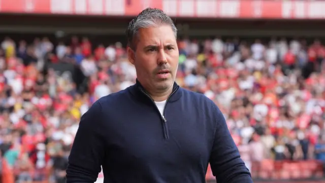 Marti Cifuentes looks ahead with a determined and assured expression on his face. He wears a navy quarter zip top and there's a crowd of people inside a stadium in the background.