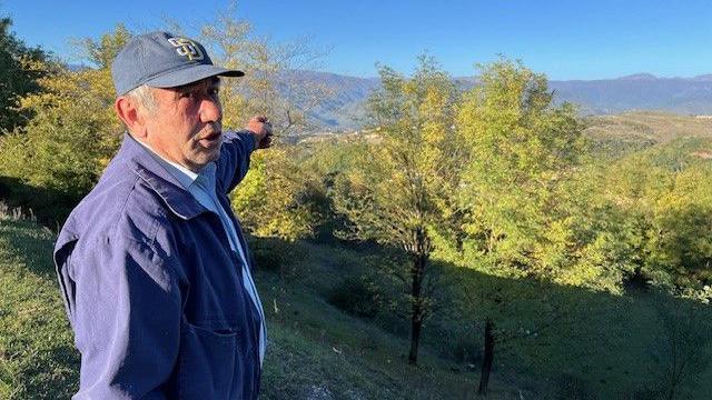 Tamaz Neparidze stands wearing a navy cap on a beautiful hillside pointing towards some trees down into a valley