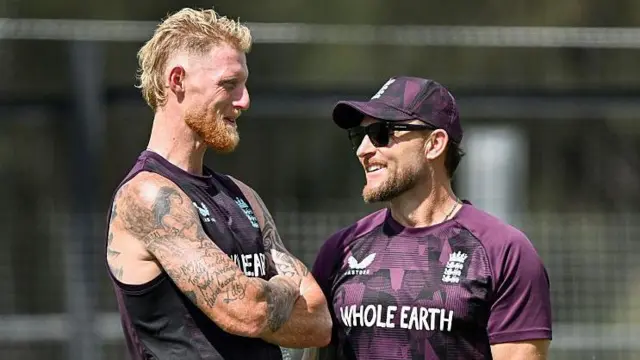 Ben Stokes (left) with his arms folded in conversation with Brendon McCullum, who is wearing a cap and sunglasses at a training session ahead of The Ashes