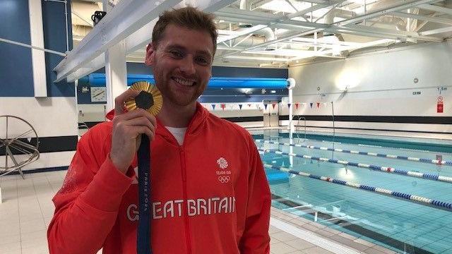 A man with blonde hair wearing a red jacket saying 'Great Britain' on it, holding up a gold medal. He's in a leisure centre and there's a swimming pool in the background.