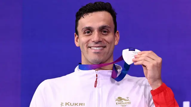 James Guy, wearing England's white and red swimming training jacket, smiles as he holds up his Commonwealth Games silver medal for the men's 200m freestyle